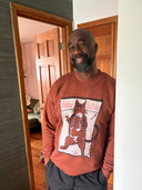 A smiling man wearing a brown crewneck sweatshirt with a graphic of the psychic sister Cat Cuddle on the front, paired with dark gray sweatpants, standing in a doorway of a home.