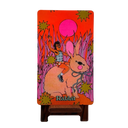 A vertical art print or decorative panel on a small wooden easel stand, featuring a colorful illustration of a child riding a rabbit against a bright orange-red background with pink and yellow starburst shapes. The word 'Rabbit' appears at the bottom.
