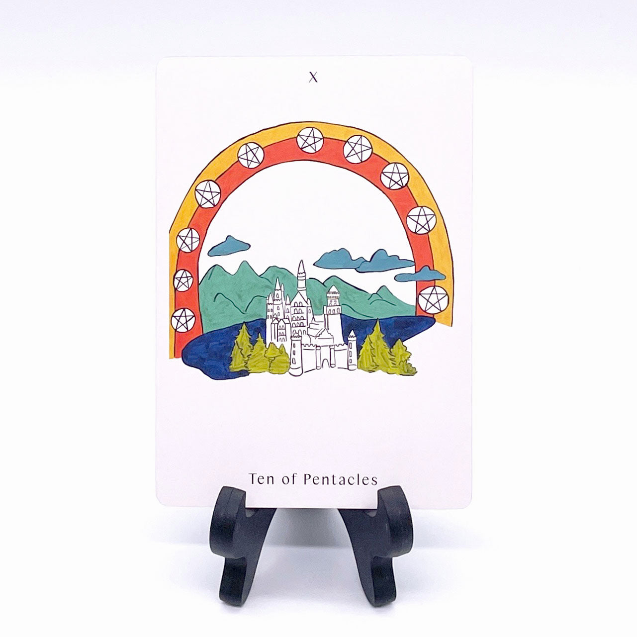 Ten of Pentacles card showing a castle on a lake against a mountain range, with an orange and red color arch over with the pentacles symbols placed across it. Card is printed with the title and corresponding number "X".